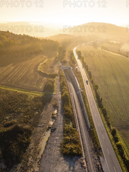 Luftaufnahme einer Landschaft im Sonnenlicht mit Eisenbahnlinie und Straße, umgeben von Feldern und Hügeln, Neubautunnel der Hermann Hessebahn, Calw, Schwarzwald, Deutschland