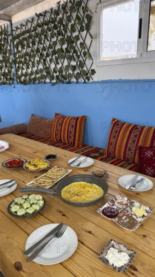 Rustic wooden table features a spread of moroccan breakfast food, with a pan of omelette, flatbread, fresh vegetables, various spreads, and vibrant textiles on the seating area