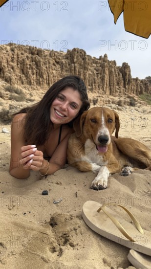 Young woman and her furry companion smiling, lying on sandy beach under an umbrella, enjoying a sunny day outdoors with eroded cliffs in the background