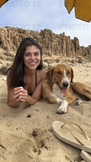 Young smiling woman with dark hair lying on a sandy beach next to a happy brown dog, both relaxing under a parasol with rocky cliffs in the background