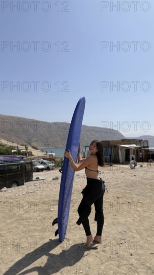 Young woman smiling, holding a blue surfboard on a dusty beach, wearing a wetsuit top and bottom, with mountains, water, and surf shacks in the background on a sunny day in morocco