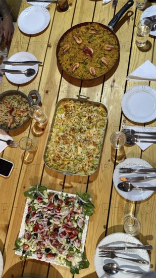 Table set for a dinner party with a large paella of shrimp, baked vegetable gratin and a vibrant salad on a wooden table, ready for sharing with family and friends