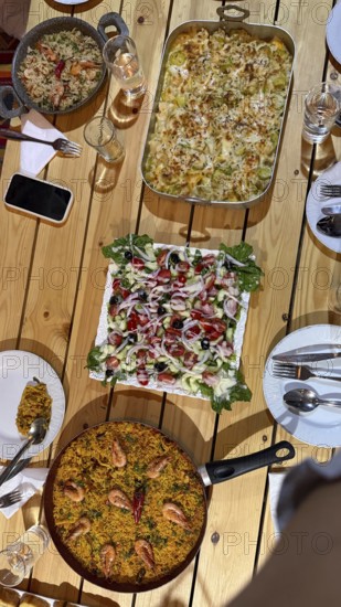 Rustic wooden table set with multiple homemade dishes including a large pan of seafood paella, a casserole, and a fresh salad, creating a vibrant feast for sharing during a meal