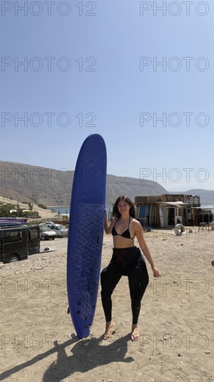 Young woman on a sunny moroccan beach holding a blue surfboard, wearing a bikini top and wetsuit bottoms, smiling and preparing to surf in calm ocean waters near taghazout
