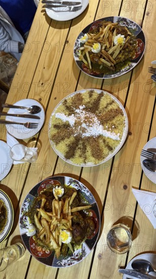 Table spread of moroccan dishes for sharing, chicken tajine topped with fries and eggs, sweet seffa medfouna dusted with powdered sugar and cinnamon, ready for a festive meal