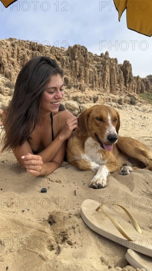 Woman lying on sand, smiling and gently petting a dog, relaxing on a beach with cliff formations in the background under an umbrella, enjoying a happy vacation moment outdoors