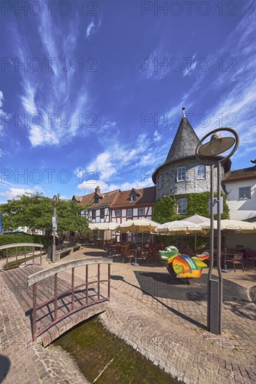 Altstadt, historische Fachwerkhäuser, Häuserzeile, hölzerne Fußgängerbrücke, Laterne, Turm, Gehweg aus Pflastersteinen und Betonpflastersteinen, Restaurant Zum Türmchen, Außenbereich einer Gastronomie mit Sonnenschirmen, Bäume, Hecke, blauer Himmel, Cumuluswolken, Cirrostratuswolken, Platz am Untertor, Burggrabenstraße, Hofheim am Taunus, Main-Taunus-Kreis, Hessen, Deutschland
