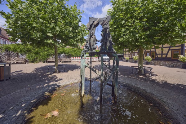 Brunnen mit Metallskulpturen, Alltägliches Theater, Bildhauer Hugo Uhl, Platz, Bäume, Hecke, schattig, blauer Himmel, Cirrostratuswolken, Kreuzung zwischen Tivertonplatz, Burgstraße, Burggrabenstraße und Taubengasse, Hofheim am Taunus, Main-Taunus-Kreis, Hessen, Deutschland