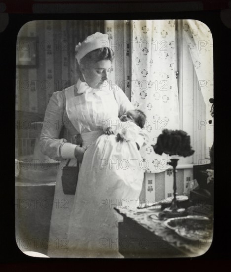 Magic lantern slide of nurse in uniform holding newborn baby inside a home, England, UK c 1900-1910