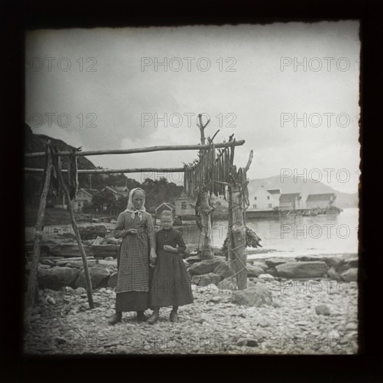 Magic lantern slide of mother and daughter standing by wooden frame of cod fish drying with village in background, Norway, c 1900