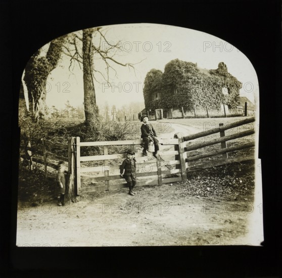 Magic lantern slide of children by gate entrance with mysterious looking ivy covered house standing on smack hill in field behind, England, UK c 1900- 1910
