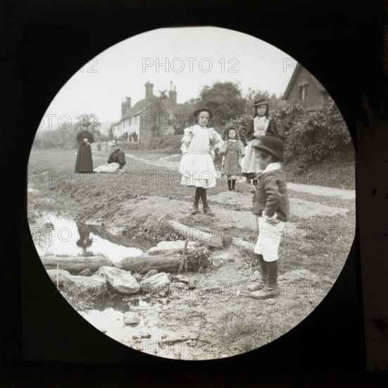 Magic lantern slide of mothers with children outside playing by a stream in a village rural location, UK circa 1900