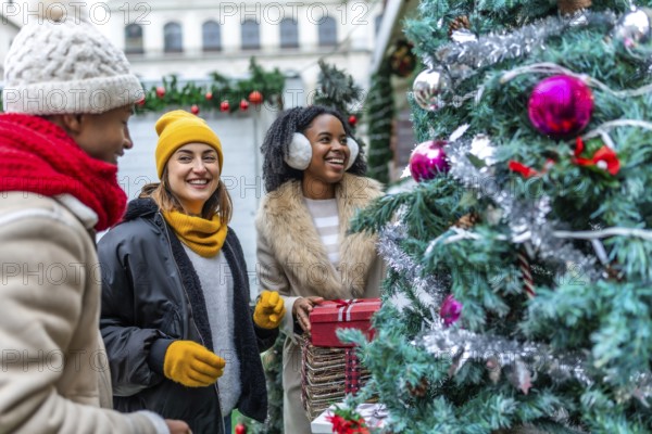 Three happy friends enjoying christmas shopping at an outdoor market, choosing a christmas tree and holding a gift box