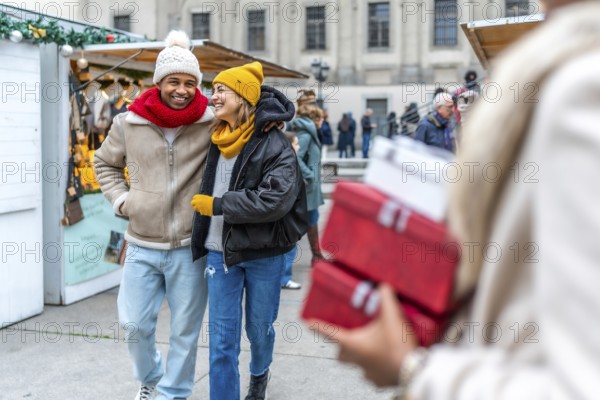 Happy multi ethnic couple walking arm in arm between christmas market stalls, woman carrying presents