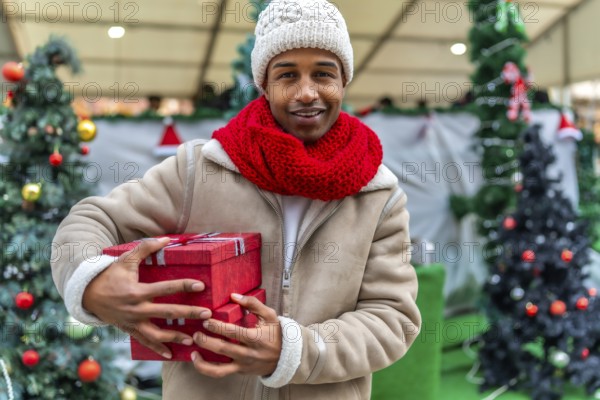 Happy young man holding colorful christmas gifts while enjoying the festive atmosphere of a bustling winter market, dressed warmly for the season