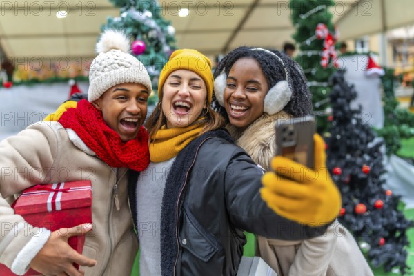 Three cheerful multi ethnic friends taking a selfie at a christmas market while holding a gift, enjoying winter holidays together