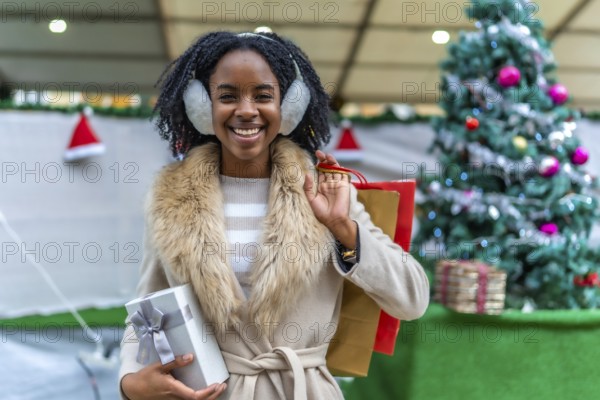 Young black woman wearing earmuffs holding christmas present and shopping bags, smiling in front of christmas tree