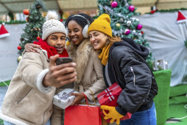 Three happy friends taking a selfie with smartphone at the christmas market, holding gifts and shopping bags