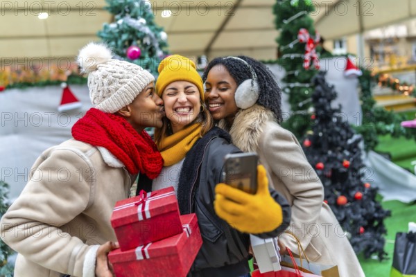 Three happy friends are taking a selfie at a christmas market, one of them is kissing another one on the cheek while holding christmas gifts
