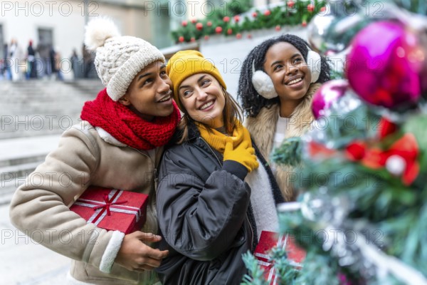 Three friends holding christmas gifts are admiring a decorated christmas tree at a city market during wintertime