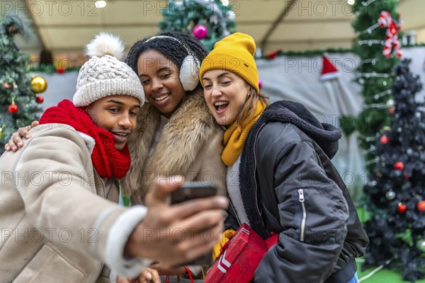 Three cheerful young friends taking a selfie with a smartphone in a city during christmas time, with winter clothes and decorations