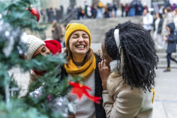 Three female friends are enjoying the christmas spirit, laughing near a decorated christmas tree in a city during winter