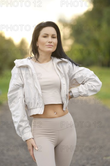 Confidently posing in a scenic park, a fitness enthusiast showcases her athletic style in a light jacket and leggings. The warm glow of sunset creates a picturesque backdrop for her workout attire