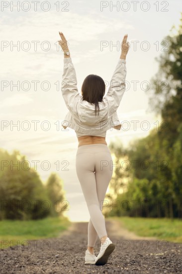 Rear view of a woman standing with her arms raised on a trail, enjoying the tranquility of nature in an outdoor environment. The soft glow of the sunset illuminates the path surrounded by lush trees