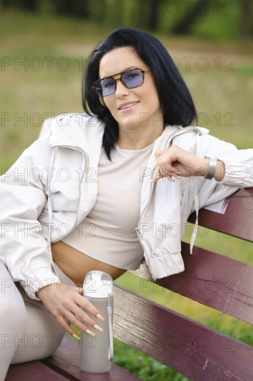 A playful woman wearing sunglasses and a light jacket is sitting comfortably on a bench in a park. She exudes a relaxed vibe, holding a drink and enjoying the warmth of a sunny day
