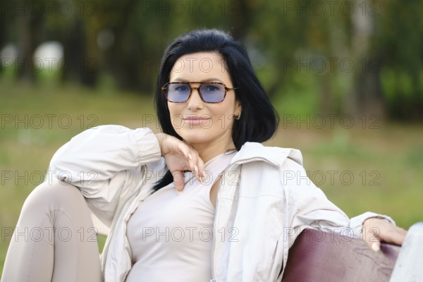 Cheerful woman enjoys a peaceful afternoon in a park, wearing sunglasses and a light jacket. She poses relaxed on a bench, surrounded by greenery, highlighting a tranquil moment outdoors