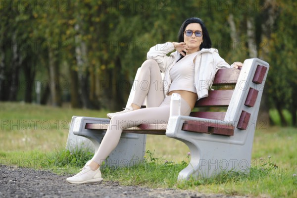 A woman sits on a park bench dressed in light-colored casual clothes, sunglasses on, with a relaxed pose