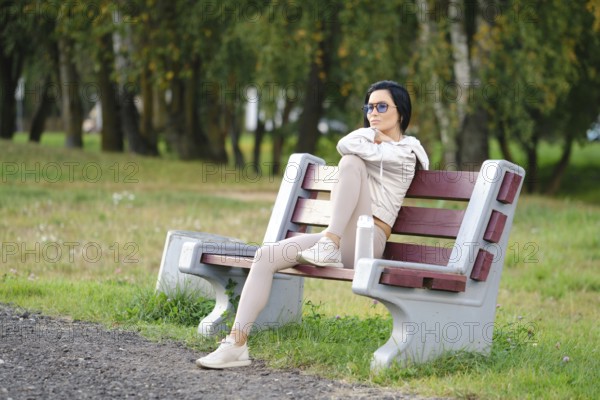 A woman sits on a park bench, wearing leggings and a light jacket. She enjoys the warmth of calm sunny evening and is deep in thought