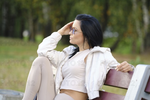 A woman with long dark hair sits on a park bench, dressed in comfortable attire. She gazes thoughtfully into the distance, surrounded by lush greenery and soft sunlight filtering through trees