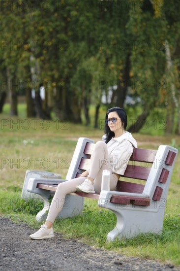 A slender woman sits comfortably on a park bench, wearing glasses and casual attire. Sunlight filters through the trees in the background, creating a serene atmosphere