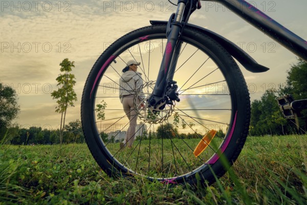 A woman enjoying a peaceful moment of sunset in nature while standing near her bicycle. She is relaxed, wearing casual attire and a cap