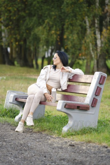 A woman dressed in comfortable casual wear sits on a park bench, gazing thoughtfully into the distance. The serene environment features trees and greenery, creating a peaceful atmosphere