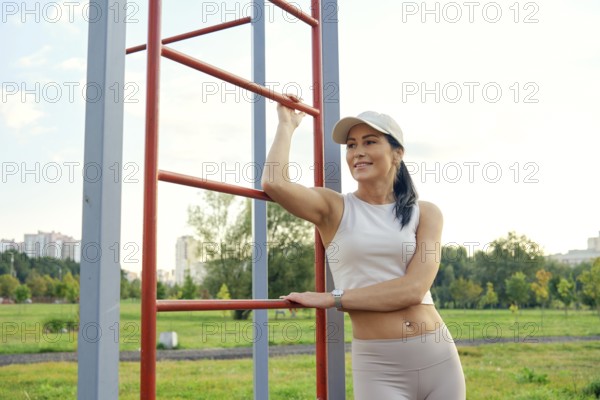 A fitness enthusiast relaxes at a park, leaning against outdoor exercise equipment. She enjoys the sunshine, showcasing her active lifestyle in a serene urban environment surrounded by greenery