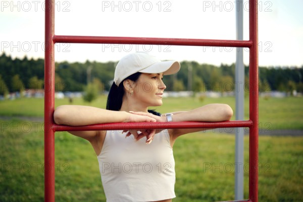 A woman in a casual outfit stands at a fitness station in a park, resting her arms on the bars while enjoying the warm afternoon sun. The greenery and trees create a vibrant backdrop