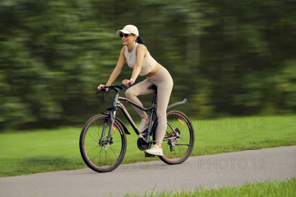 A slender woman in a tank top, cap and leggings quickly rides on a bicycle along the alley in a city park