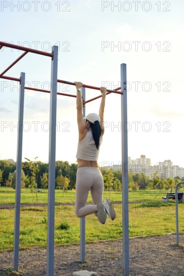 A woman is performing an exercise on pull-up bars in a city park. The sun is setting, casting a warm glow, while she focuses on her workout amidst greenery and urban buildings in the background