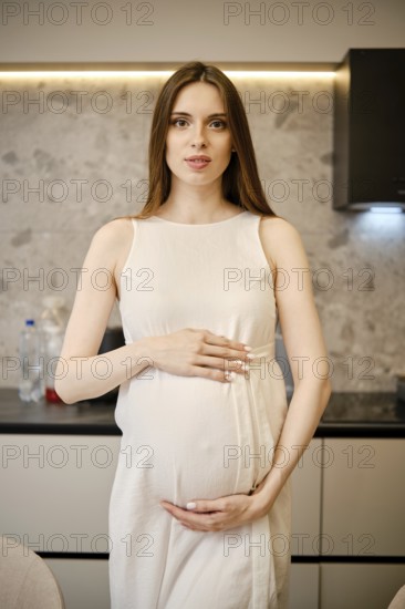 A pregnant woman stands in a contemporary kitchen, gently cradling her baby bump. She has long hair and appears calm, dressed in a simple light-colored dress, against a stylish backdrop