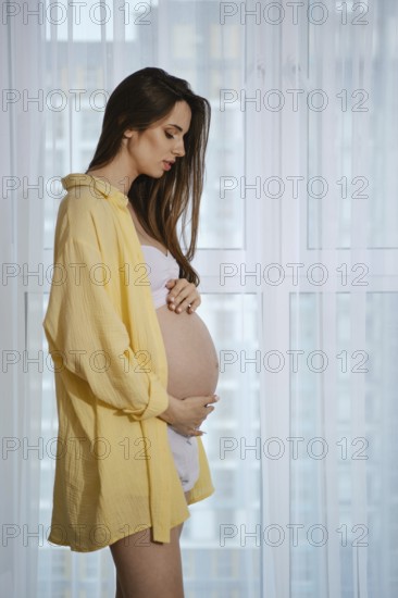 A pregnant woman stands near a window in profile, wearing a yellow shirt and white outfit, lovingly caressing her baby bump. Soft light enters the room, creating a warm and serene atmosphere