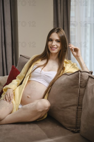 A relaxed expectant mother sits comfortably on a soft sofa, wearing a light outfit that highlights her baby bump. Sunlight filters through the curtains, creating a warm and inviting atmosphere