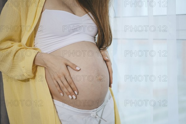 Close-up view of a baby bump of pregnant woman by the window. A woman lovingly places her hands on her well-defined baby bump, showcasing the beauty of motherhood during the morning hours