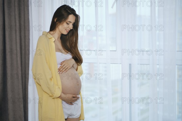 A pregnant woman stands by a window, smiling as she gently cradles her baby bump. Soft morning light filters through sheer curtains, creating a warm and serene atmosphere in the room