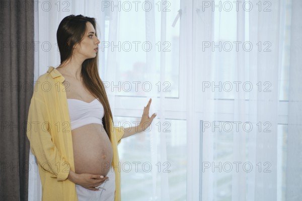A pregnant woman stands by a window in a contemporary apartment, wearing a white top and loose yellow shirt. She gently touches her belly and gazes thoughtfully outside, basking in the natural light