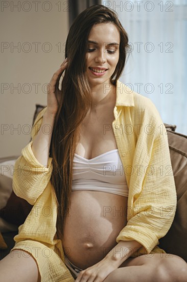 A pregnant woman in a yellow shirt and white top, sitting comfortably at home. She touches her hair and smiles, enjoying the tranquility of her surroundings