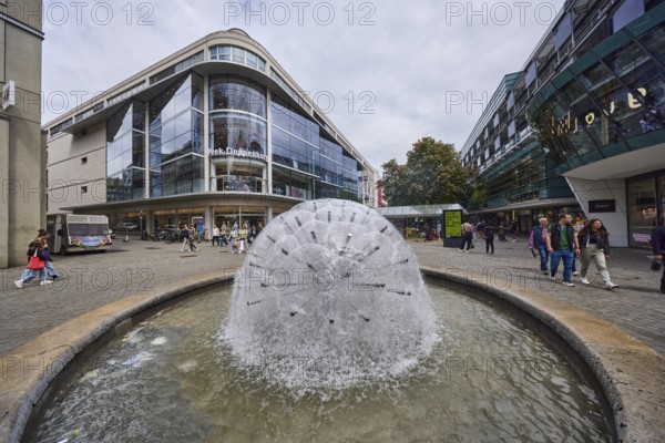Dandelion fountain, stainless steel fountain, building, pedestrian zone, retail shops, shop window, pedestrians as accessories, trees, milky blue sky, diffuse light, slightly sunny, intersection Königstraße with Thouretstraße, Stuttgart, state capital, city district Stuttgart, Baden-Württemberg, Germany