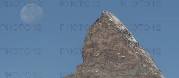 The Matterhorn mountain peak rises steeply against a bright blue sky. The full moon glistens gently above, creating a marvellous view in the serene evening hours. Matterhorn, Zermatt, Valais, Alps, Swiss