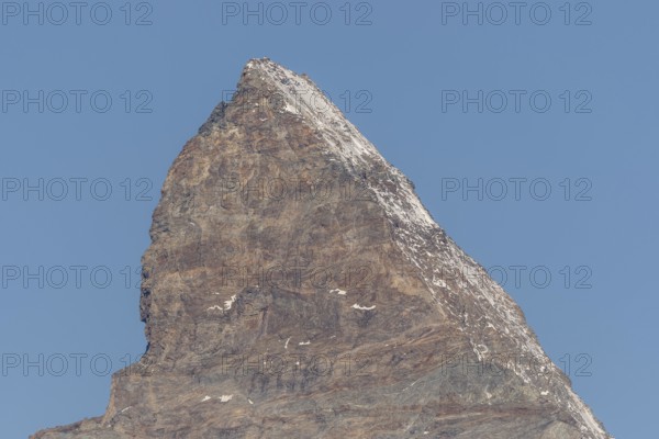 A towering Matterhorn mountain peak stands prominently against the clear blue sky, revealing rocky slopes and a thin blanket of snow on its summit during the bright midday sun. Zermatt, Valais, Alps, Swiss